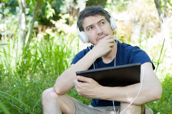 Młody Człowiek Relaksujący Tabliczka Słuchania Muzyki Słuchawkami Uszach Parku Odkryty — Zdjęcie stockowe