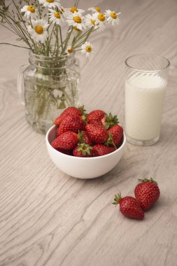 Çilekli beyaz kase ve papatya buketli bir bardak süt.