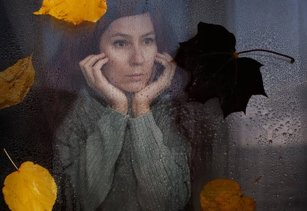 the girl looks out the window at the rain and autumn foliage