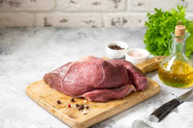 Açık gri mutfak masasındaki ahşap bir tahtada jambon eti. Çiğ et. Büyük bir parça et, tuz, biber ve sebze yağı kaselerinin yanında, kesim tahtasının üzerinde duruyor.