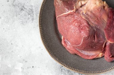 Çiğ bir sığır bacağı açık gri mutfak masasında seramik bir tabakta oturur. Et eti. Büyük bir et parçası bir tabak içinde, bir kase biber ve bir bıçağın yanında yatıyor. Yukarıdan görünüşü