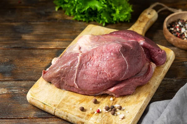 Beef ham on a wooden Board on a wooden kitchen table. Raw beef meat. A large piece of meat lies on the cutting Board, next to a wooden bowl with salt and pepper
