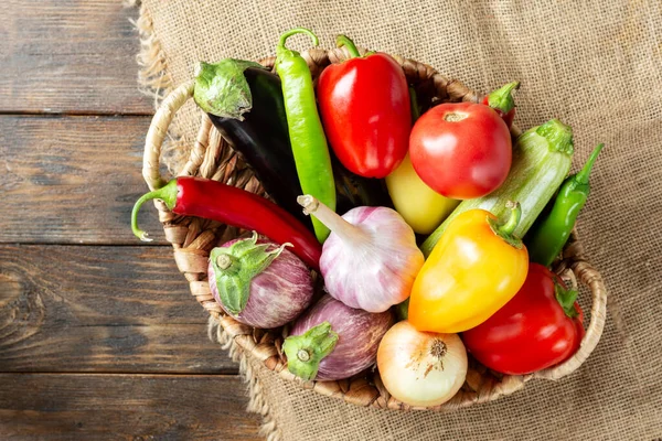 Kahverengi ahşap bir masada sepetteki taze sebzeler. Sepette bir sürü çiğ sebze var. Patlıcan, domates, sarımsak, tatlı biber, masada soğan. Metin için alanı olan üst görünüm. Sağlıklı gıda