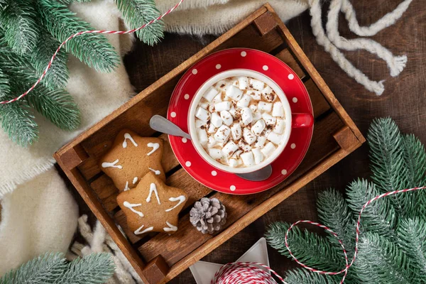 Cocoa with marshmallows in a red Cup and gingerbread on a tray on a Christmas background. Red decorations on spruce branches on a brown background. Christmas Flatly. Top view