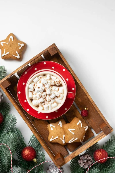Cocoa with marshmallows in a red Cup on a tray on a white Christmas background. Red decorations on spruce branches on a white background. Christmas Flatly. Top view from copyspace