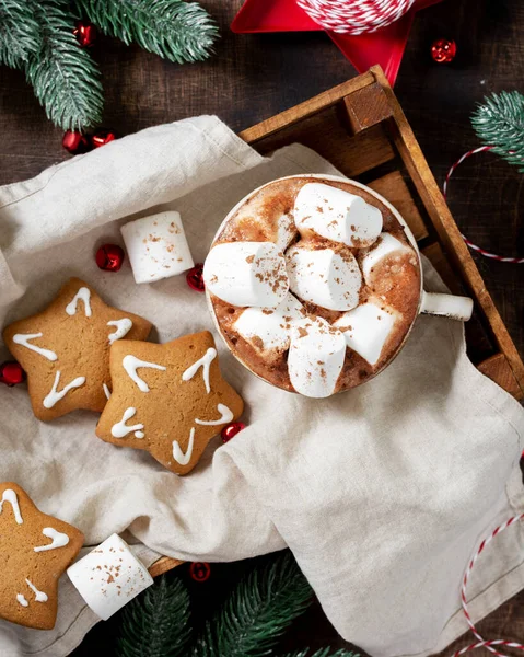 Hot cocoa or hot chocolate in a ceramic Cup on a wooden tray surrounded by gingerbread, Christmas tree branches and ornaments on a brown wooden table. Christmas Flatly