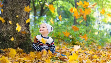 Çocuk bir ağacın altında oturur ve düşen yapraklara bakar.