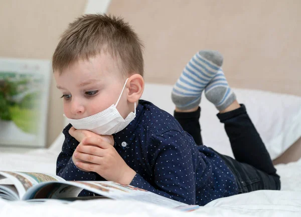 baby in a medical mask on a sofa reading a book