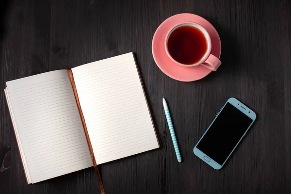Libro abierto, taza de té, bolígrafo, teléfono sobre fondo negro — Foto de Stock