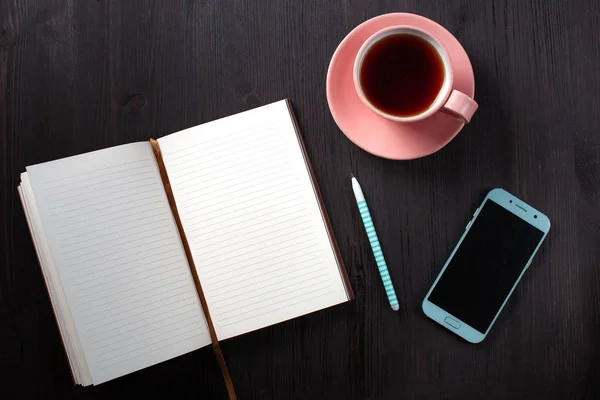 Notebook aberto, xícara de chá, caneta, telefone no fundo preto — Fotografia de Stock