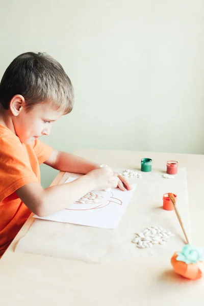 7 tahun anak laki-laki tua membuat labu dari biji di atas kertas putih duduk di meja — Stok Foto