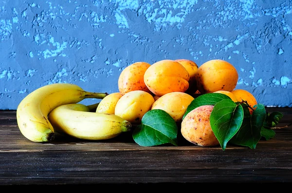 Спелые абрикосы и бананы с зелеными листьями на деревянном столе — стоковое фото
