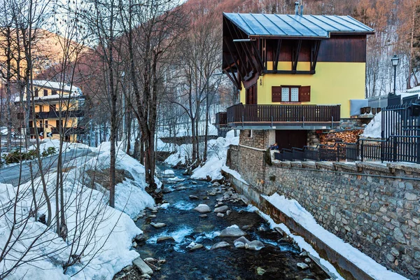 Il torrente di montagna scorre lungo una riva innevata e una casa in legno . — Foto Stock