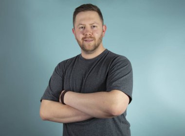 Sakallı rahat genç adam onun fotoğraf makinesi ile çapraz silah ver frontal stüdyo portre ikindilerde bakarak bir gri t-shirt