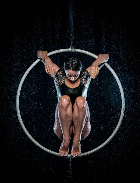Beautiful female acrobat sitting on symmetric pose in aerial hoop under rain on black background.