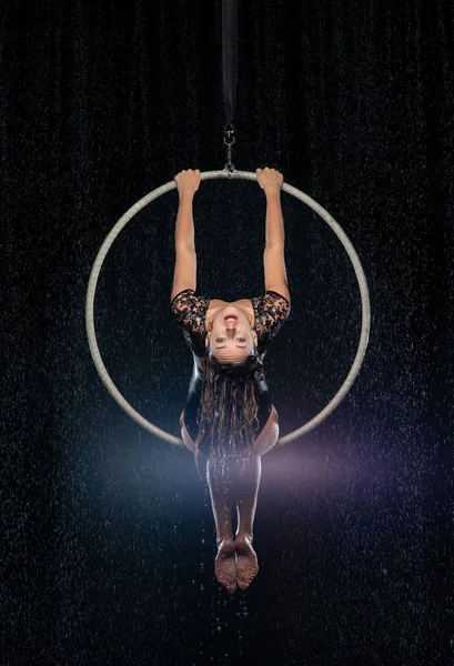 Beautiful female acrobat sitting on symmetric pose in aerial hoop under rain on black background.