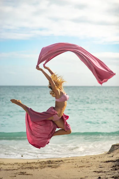 Concept de liberté et de bonheur. Femme heureuse sur la plage sautant avec de la soie rose volant — Photo