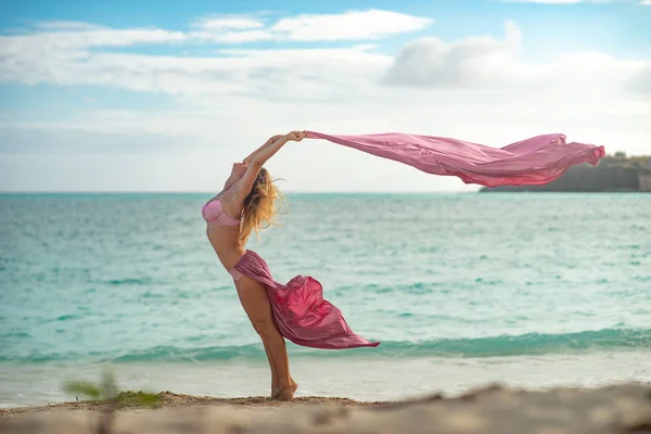 Chica joven en forma y deportivo posando en una playa con seda voladora rosa — Foto de Stock