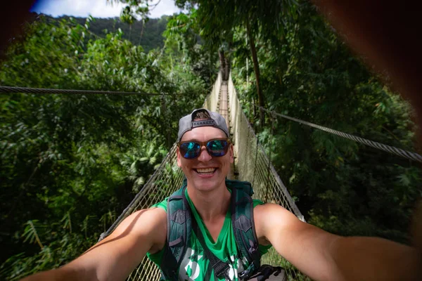 Asma köprüde gülümseyen ve özçekim yapan genç bir adam. Macera ve seyahat kavramı — Stok fotoğraf