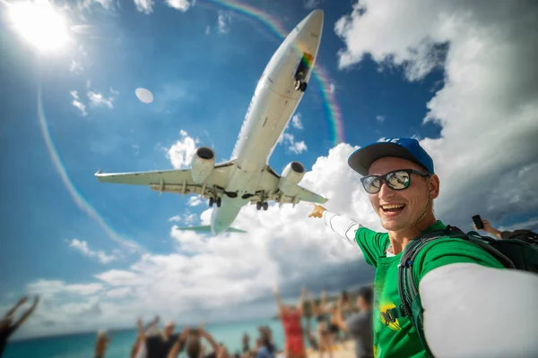 Lächelnder Mann beim Selfie mit dem Flugzeug mit Strand, Meer und vielen Touristen im Hintergrund. Maho Beach. Glücklicher Urlaub und Reisekonzept — Stockfoto