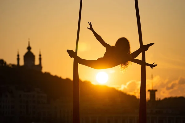 Silueta de una mujer acróbata flexible sobre seda aérea durante una puesta de sol en el fondo de la ciudad de Kiev. concepto de libertad y paz — Foto de Stock