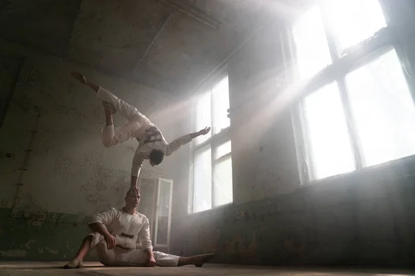 Manliga Duo gör akrobatiska tricks bär kostym av vansinniga människor i övergivna rum med solstrålar från Windows — Stockfoto