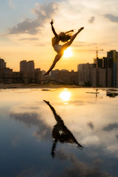 Bailarina flexible saltando al atardecer sobre fondo urbano con reflejo en el agua. Concepto de libertad y felicidad — Foto de Stock