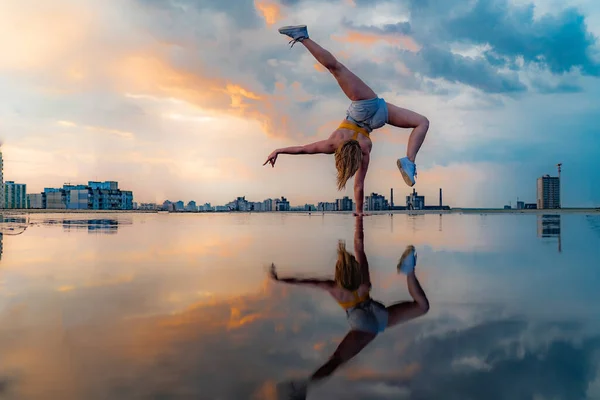 Gimnasta femenina de pie por un lado y manteniendo el equilibrio durante la dramática puesta de sol con reflejo en el agua de nubes increíbles. Concepto de Calistenia, contorsión y soporte de mano — Foto de Stock