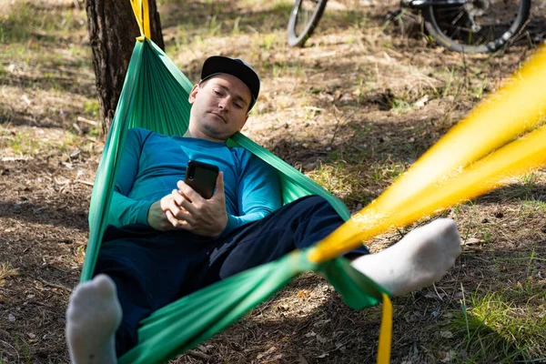 Genç ve mutlu bir adam cep telefonunu şapkasında kullanıyor. Dışarıda, ormanda, hamakta dinleniyor.. — Stok fotoğraf