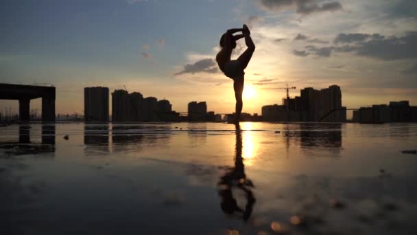 Flexible female gymnast doing acrobatic tricks with reflection in the water during dramatic sunset with cityscape background. Concept of fearless, courage and freedom — Stock Video