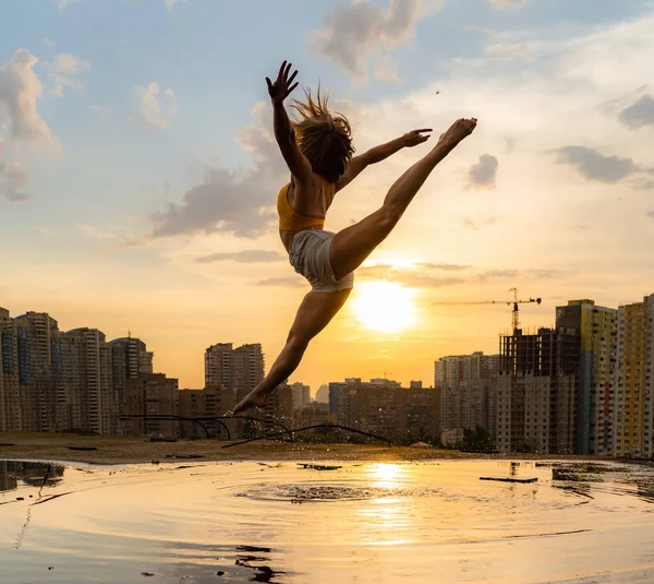 Bailarina flexible saltando al atardecer sobre fondo urbano con reflejo en el agua. Concepto de libertad y felicidad — Foto de Stock