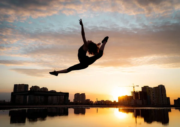Bailarina flexible saltando al atardecer sobre fondo urbano con reflejo en el agua. Concepto de libertad y felicidad — Foto de Stock
