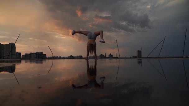 Gymnaste féminine flexible faisant handstand et calisthénique avec réflexion dans l'eau sur fond de paysage urbain pendant le coucher du soleil dramatique au ralenti — Video