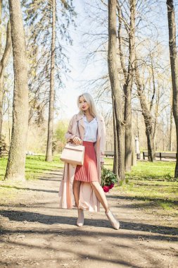 Pembe bez içinde şık kadın moda portre. İpeksi yağmurluk, ayak bileği botları, etek ve bluz holding el çantası ve çiçek kız