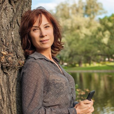 Olgun kadın açık havada dikey. Park'ta rahatlatıcı smartphone ile orta yetişkin kadın