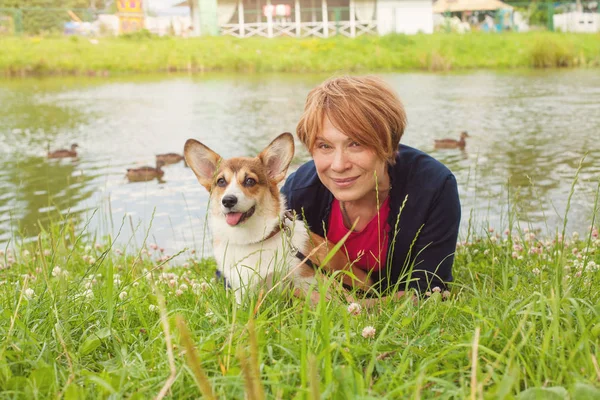 Досить старша жінка лежить на зеленій траві і обіймає свого собаку-коргі — стокове фото