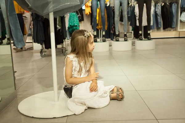 Kid meisje in witte jurk wacht mama in de winkel. Kinderen vermoeidheid — Stockfoto