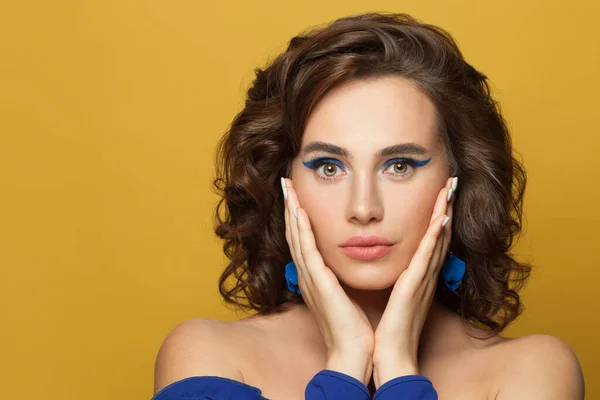 Beautiful brunette woman face. Model with brown curly hair and blue eyeliner arrow make up on vivid yellow background