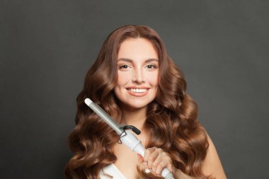 Charming woman with long wavy hairstyle using hair curling tongs iron, indoor portrait. Hairdresser equipment tools and hair styling concept