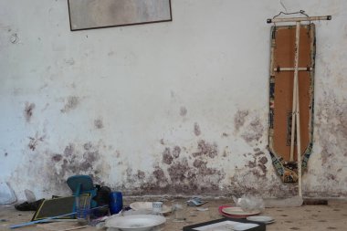 Ütü masası ve terk edilmiş evin içindeki diğer klasik ev eşyaları. Küflü duvar ve kirli zemin.