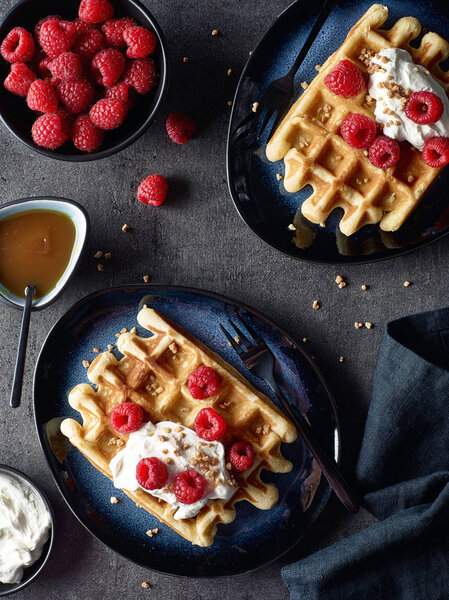 Two plates of belgian waffles with whipped cream and raspberries on dark marble background. Top view