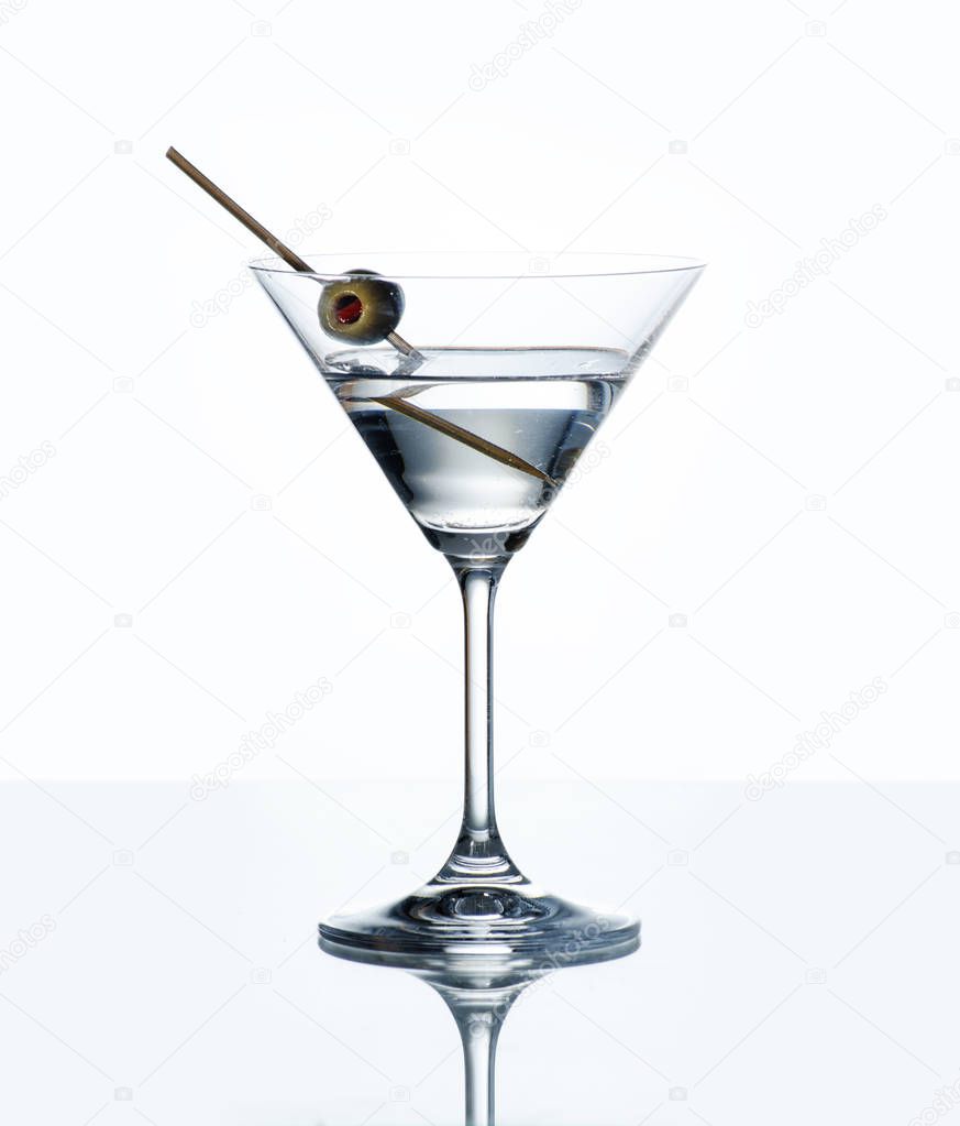 Dry martini with green olive in cocktail glass over white background with reflection. Luxury alcohol drink on bar counter.