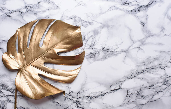 Golden monstera leaf on luxury marble background. Clean minimal flat lay design. Tropical gold foliage for trendy layout. Luxury fashion lifestyle concept.