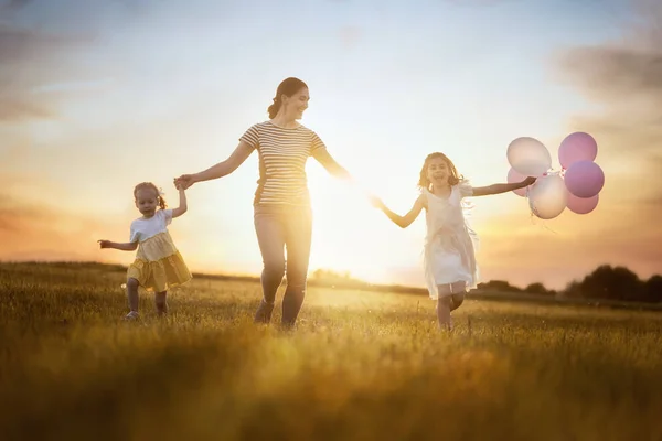 在夏天在性质上开心快乐充满爱的家庭 年轻的母亲和两个女儿都是笑着 在日落背景的草地上嬉戏 — 图库照片