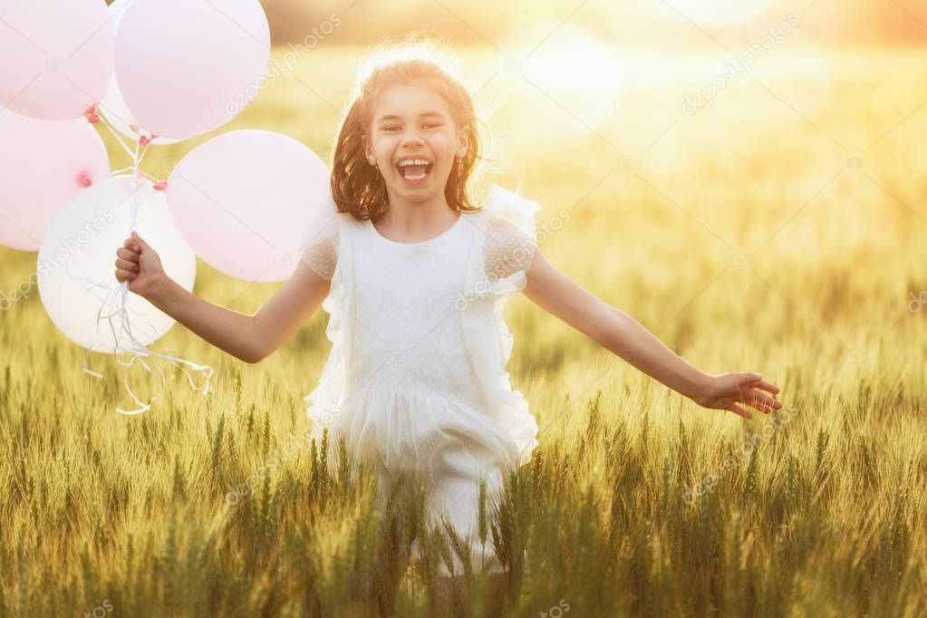 Happy kid is having fun on nature in the summer. Child is laughing and playing on meadow at sunset background. Girl with air balloons is running on cereal field.