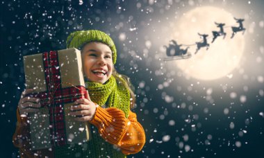 Mutlu Noeller! Xmas hediyesi ile şirin küçük çocuk. Noel Baba onun kızak ay gökyüzü uçan. Mutlu tatil keyfini çıkarın. Koyu arka plan üzerinde hediyelerle kız portresi.