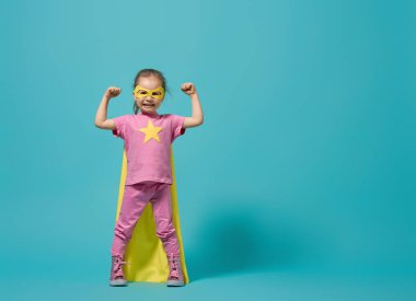 Küçük çocuk süper kahramanı oynuyor. Parlak mavi duvarın arkasındaki çocuk. Kız gücü konsepti. Sarı, pembe ve turkuaz renkler.