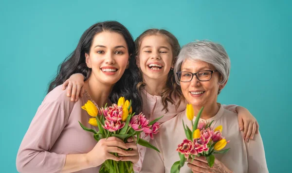 幸せな女性の日 子娘は ママとおばあちゃんの黄色の花チューリップを与えて祝福です おばあちゃん ママと女の子笑顔と光を抱いてブルーの背景 家族の休日と一体感 — ストック写真