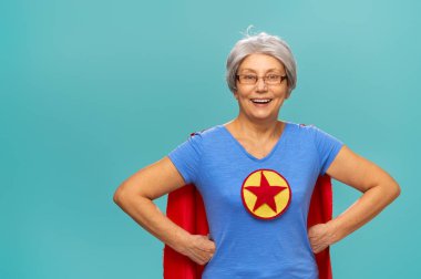 Joyful beautiful senior woman in superhero costume posing on turquoise background.