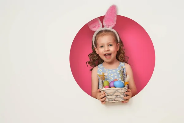 イースターの日にバニーの耳を着てかわいい小さな子供 白とピンクの背景に塗られた卵を持つ少女 — ストック写真